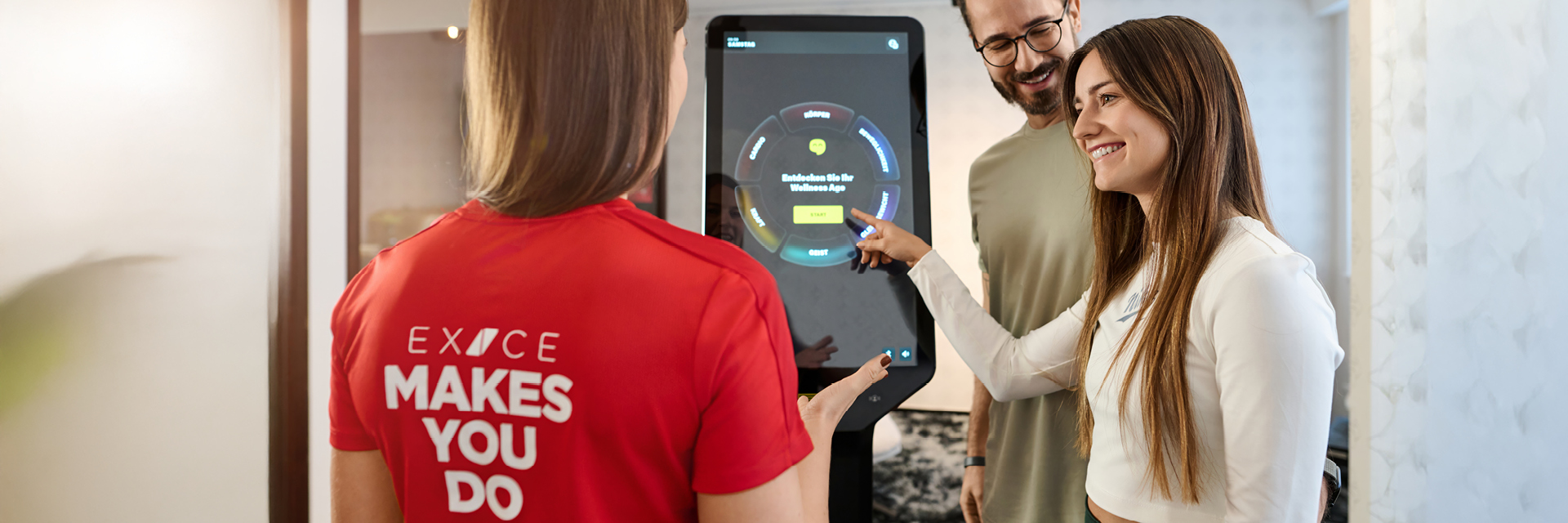 Trainerin im roten Exice-Fitness-Shirt erklärt zwei Mitgliedern an einem interaktiven Display das digitale Trainingsprogramm.